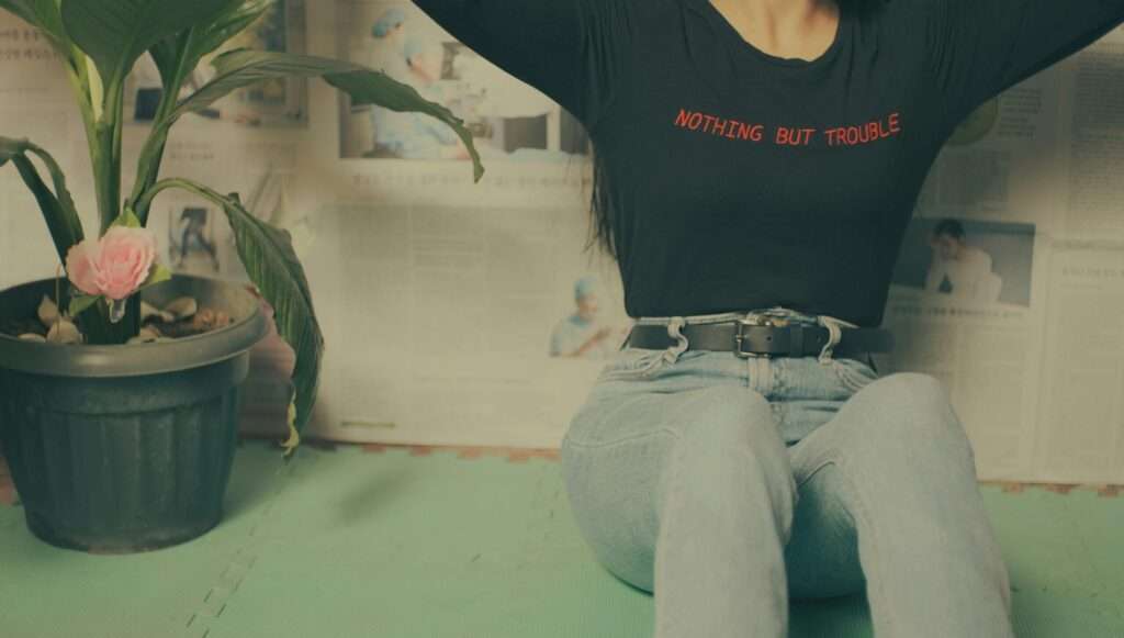 Crop anonymous female in trendy clothes sitting on floor near wall covered in newspapers near potted plant with green leaves near fake pink flower in light apartment