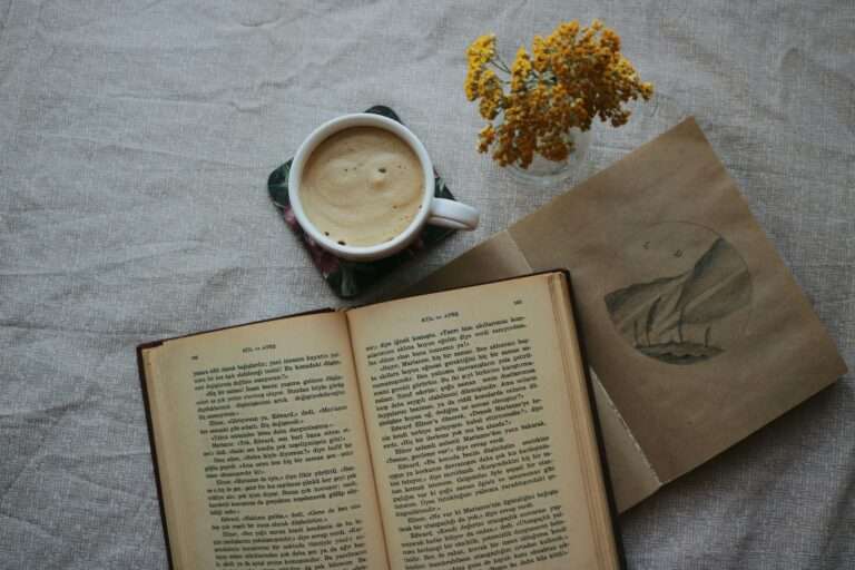 Warm overhead view of coffee, an open book, and yellow flowers on a fabric table.